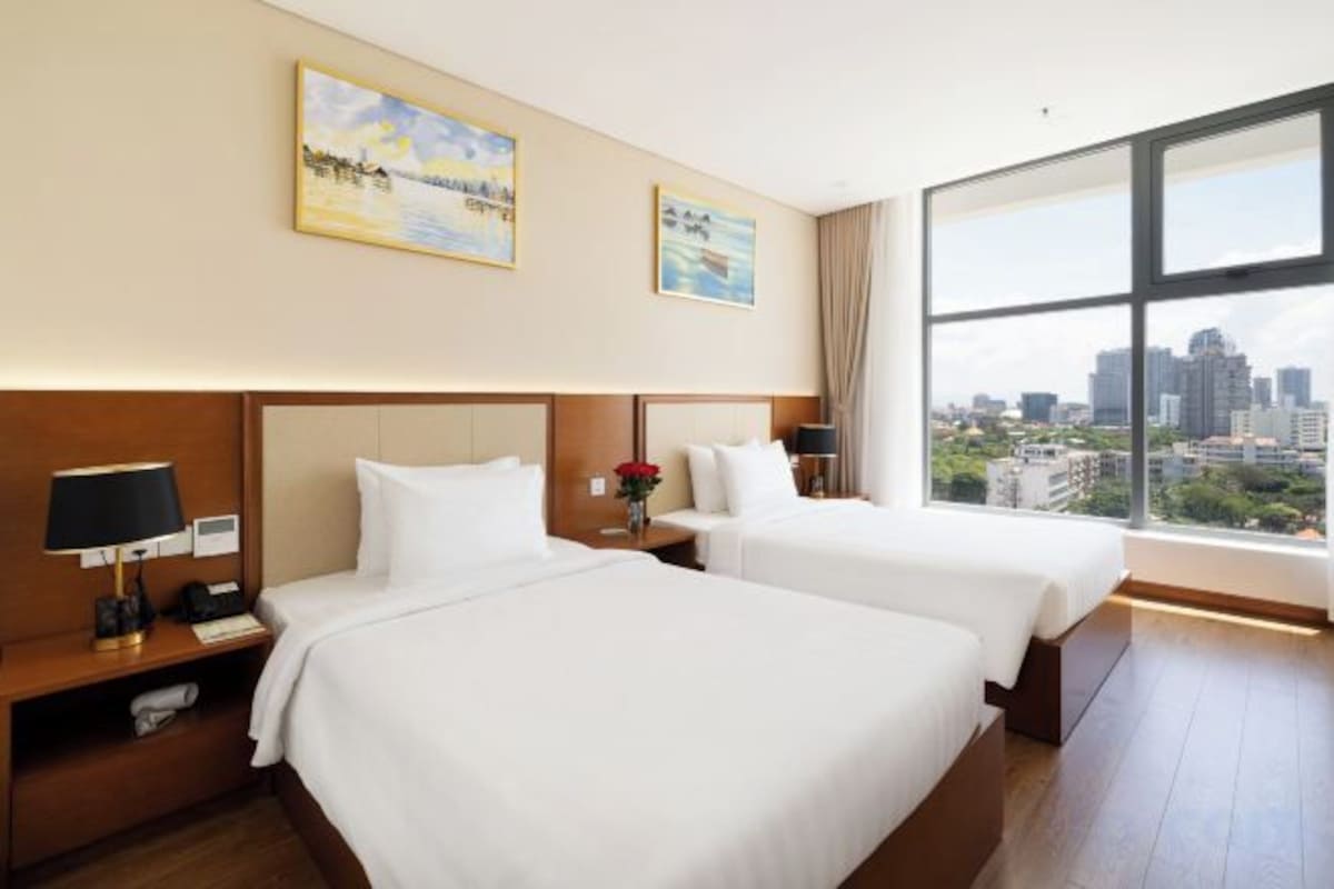 A hotel room with two beds, a large window with a city view, and a nightstand with a lamp.
