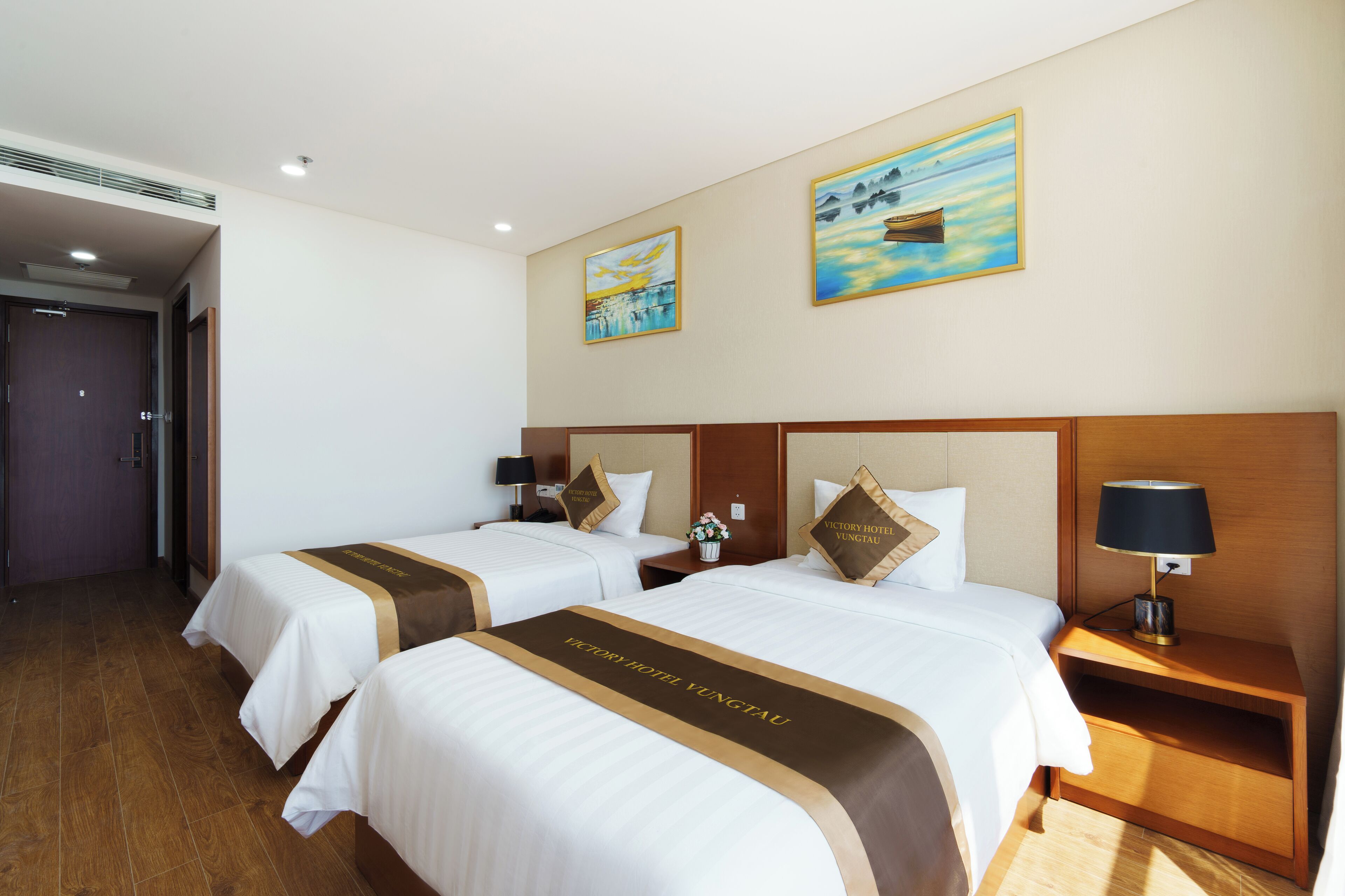 A hotel room with two beds, a nightstand, a lamp, and artwork on the walls.