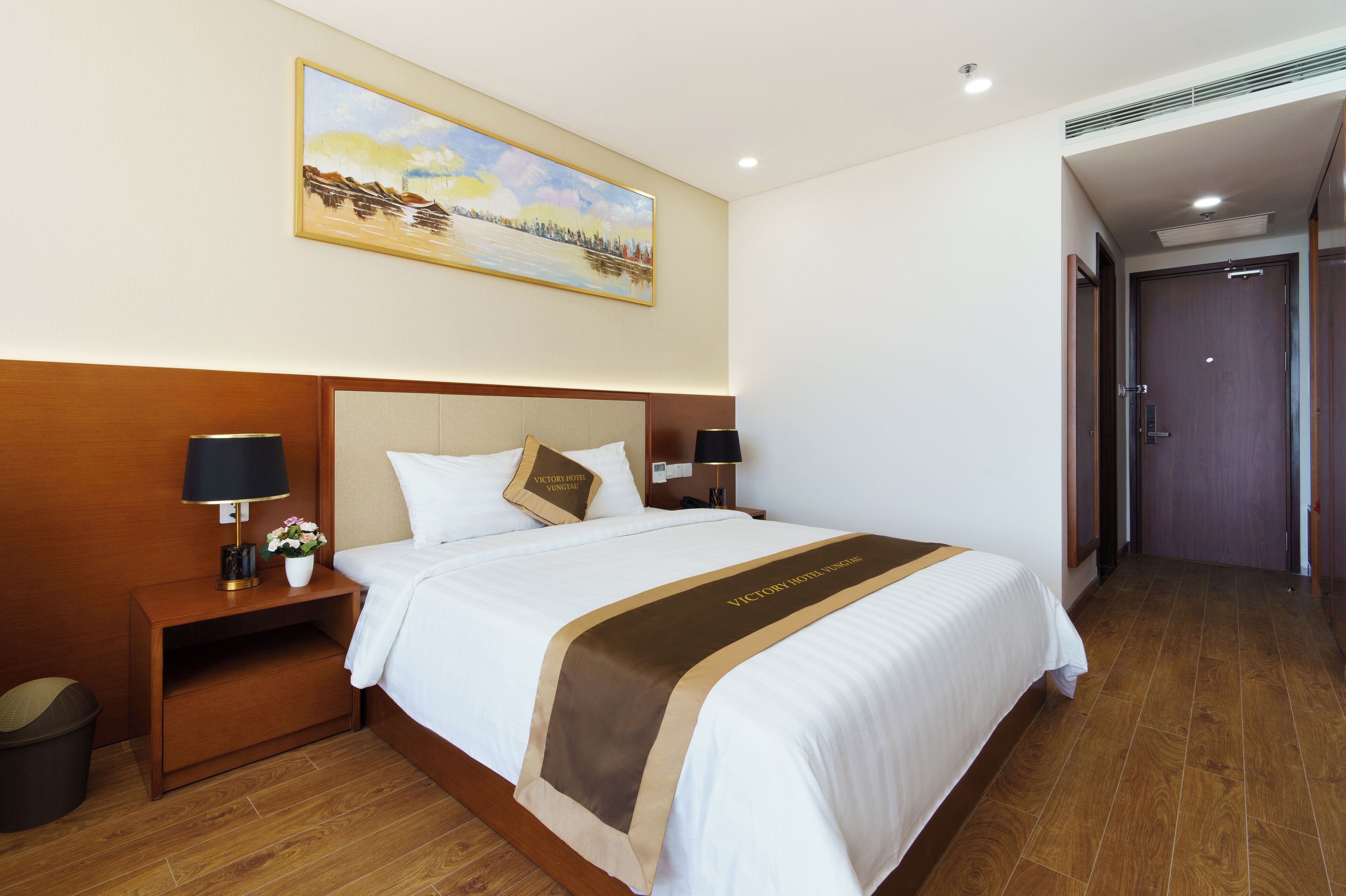 A hotel room with a large bed, a nightstand, a lamp, a painting on the wall, and a wooden floor.