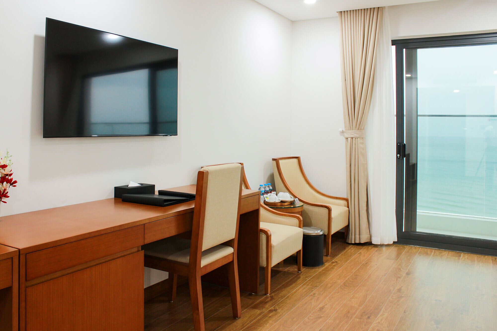 Habitación de hotel con escritorio de madera, dos sillas, televisor, una mesita con artículos de aseo y vista al mar a través de una puerta corrediza.