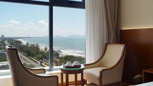 Habitación de hotel con un ventanal amplio que ofrece vistas a una ciudad costera, dos sillones tapizados, una mesita redonda con una bandeja y una cama con cabecero de madera.