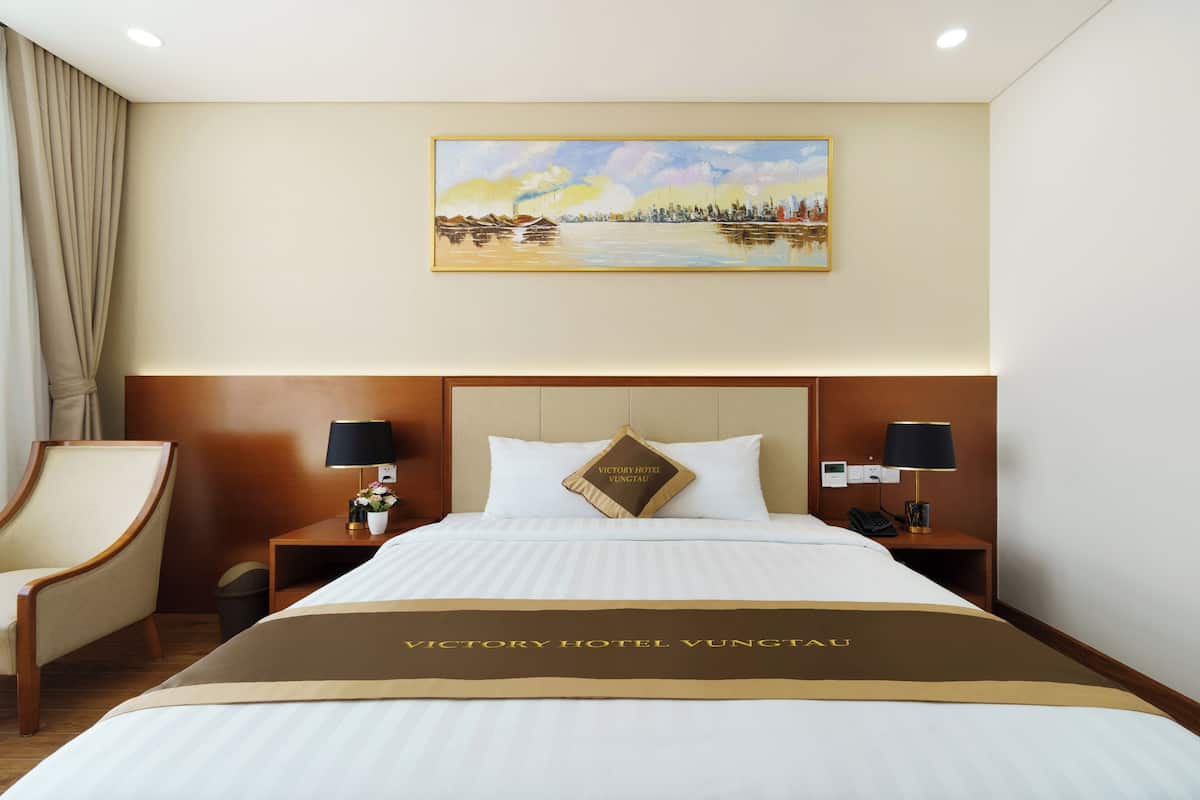 Une chambre d’hôtel avec un lit, deux tables de chevet avec des lampes, une chaise, un tableau au mur et un panneau sur la couverture du lit.