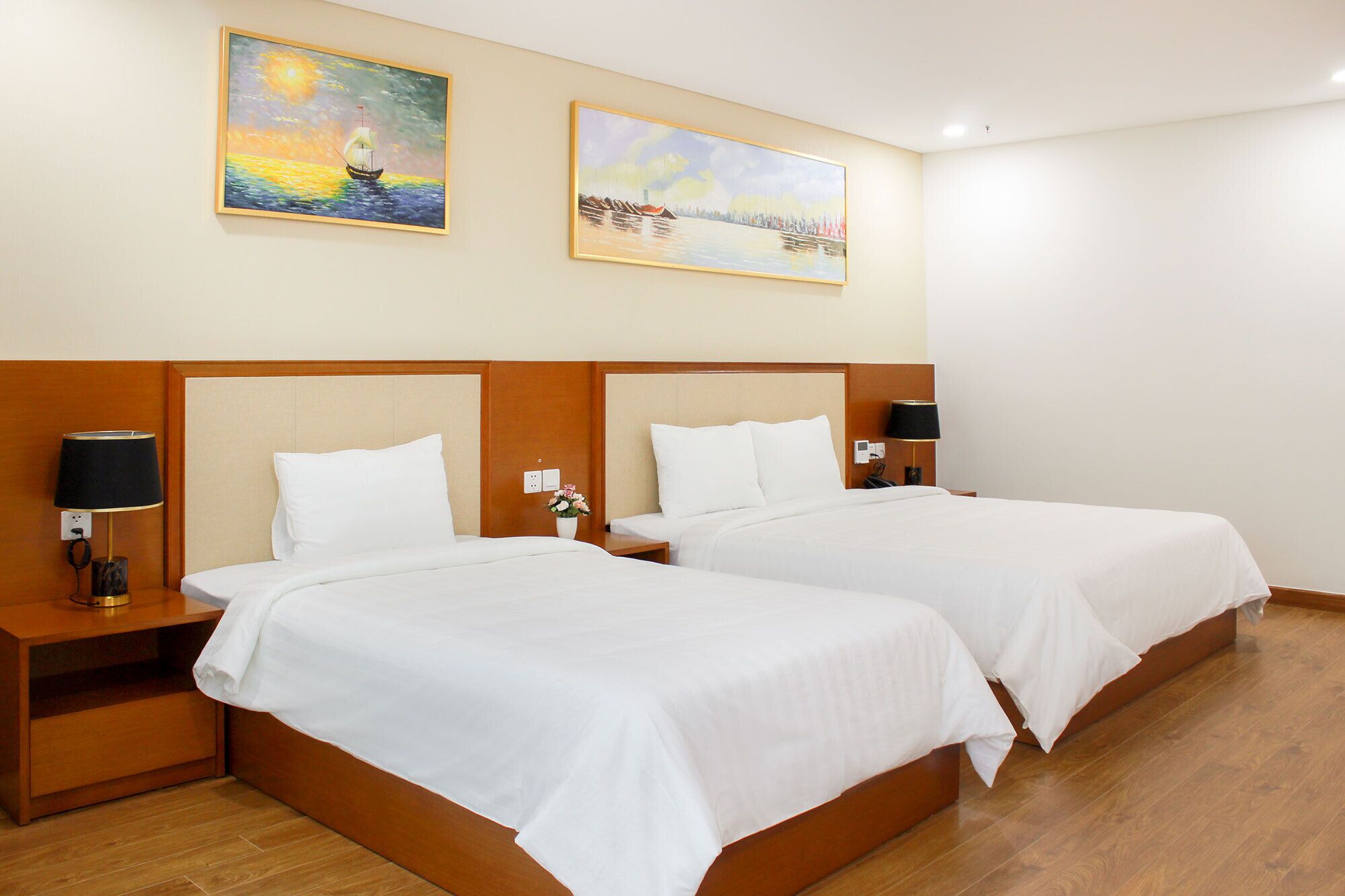 A hotel room with two beds, wooden headboards, bedside tables with lamps, and two paintings on the wall.