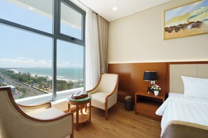 Habitación de hotel con una ventana grande, una cama, un sillón, una mesita de noche con una lámpara y un cuadro en la pared.