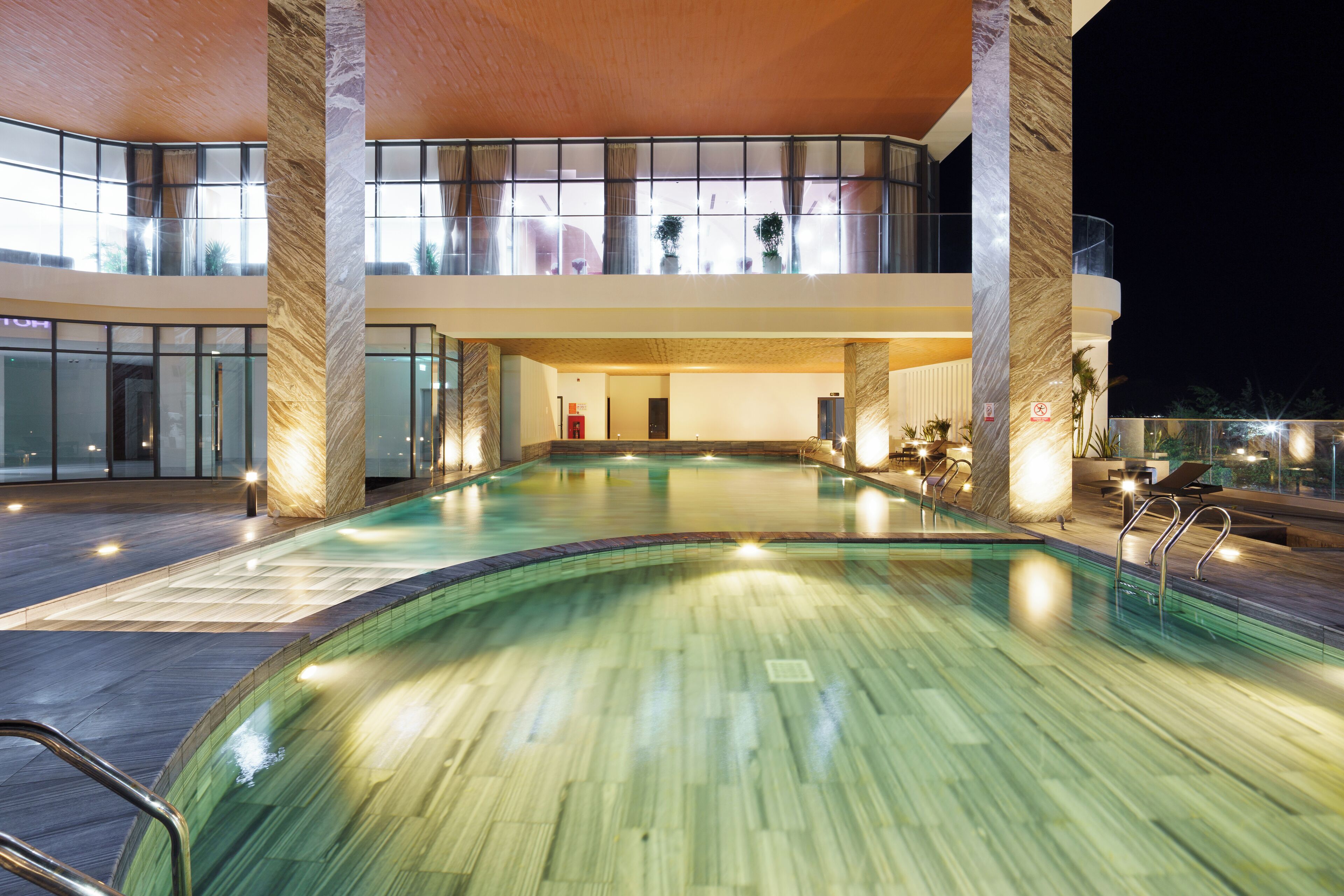 A large indoor swimming pool with a wooden floor, surrounded by glass walls and a balcony.