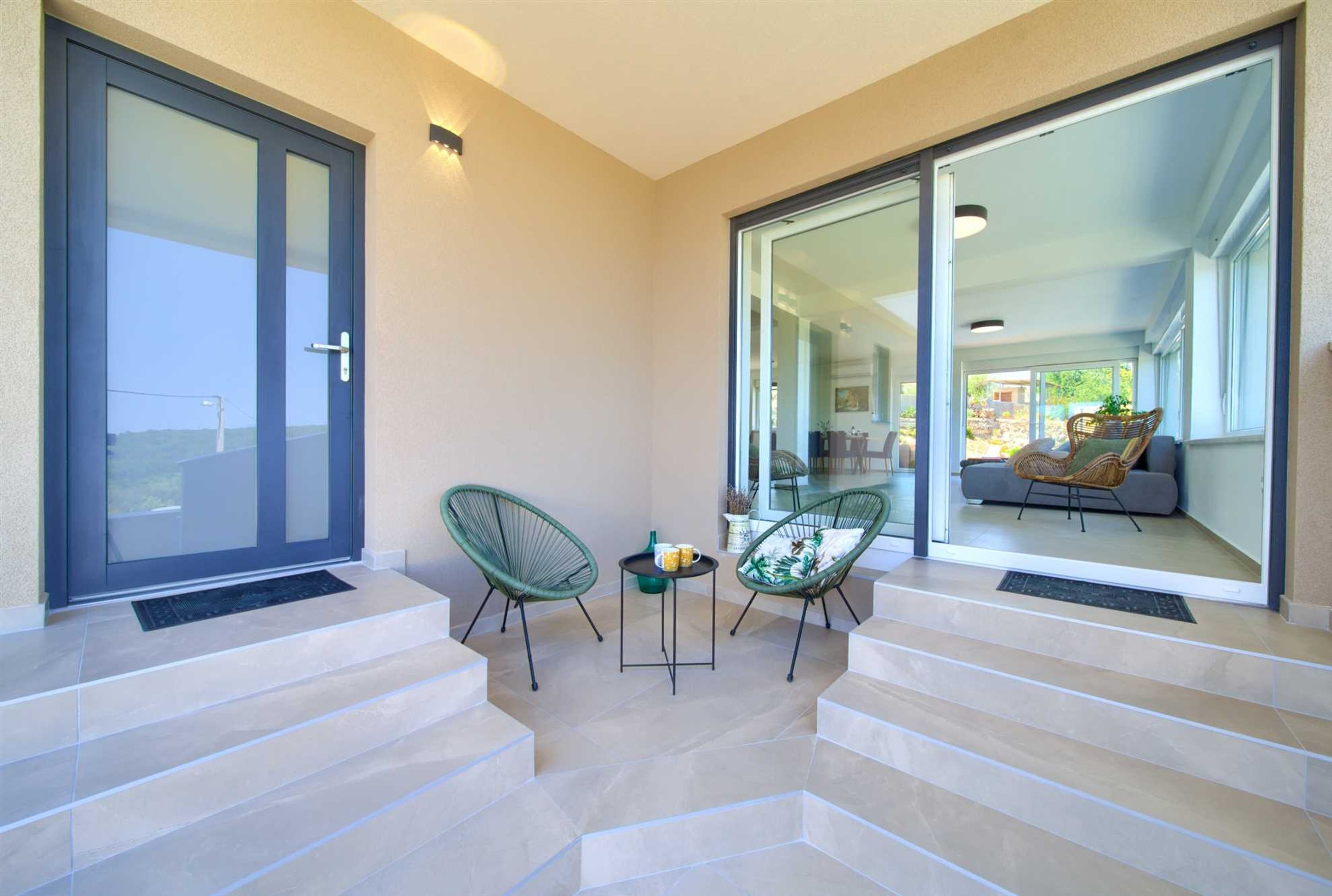 A modern outdoor area with a glass door leading to a balcony, two green chairs, and a small table with a mug.