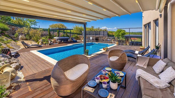 A poolside area with a wooden deck, lounge chairs, a dining table with a meal, and a view of the pool and surrounding landscape. - Villa Bella Vista in Tribulje (Dobrinj)