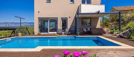 A modern two-story house with a swimming pool, outdoor seating, and a view of the mountains.