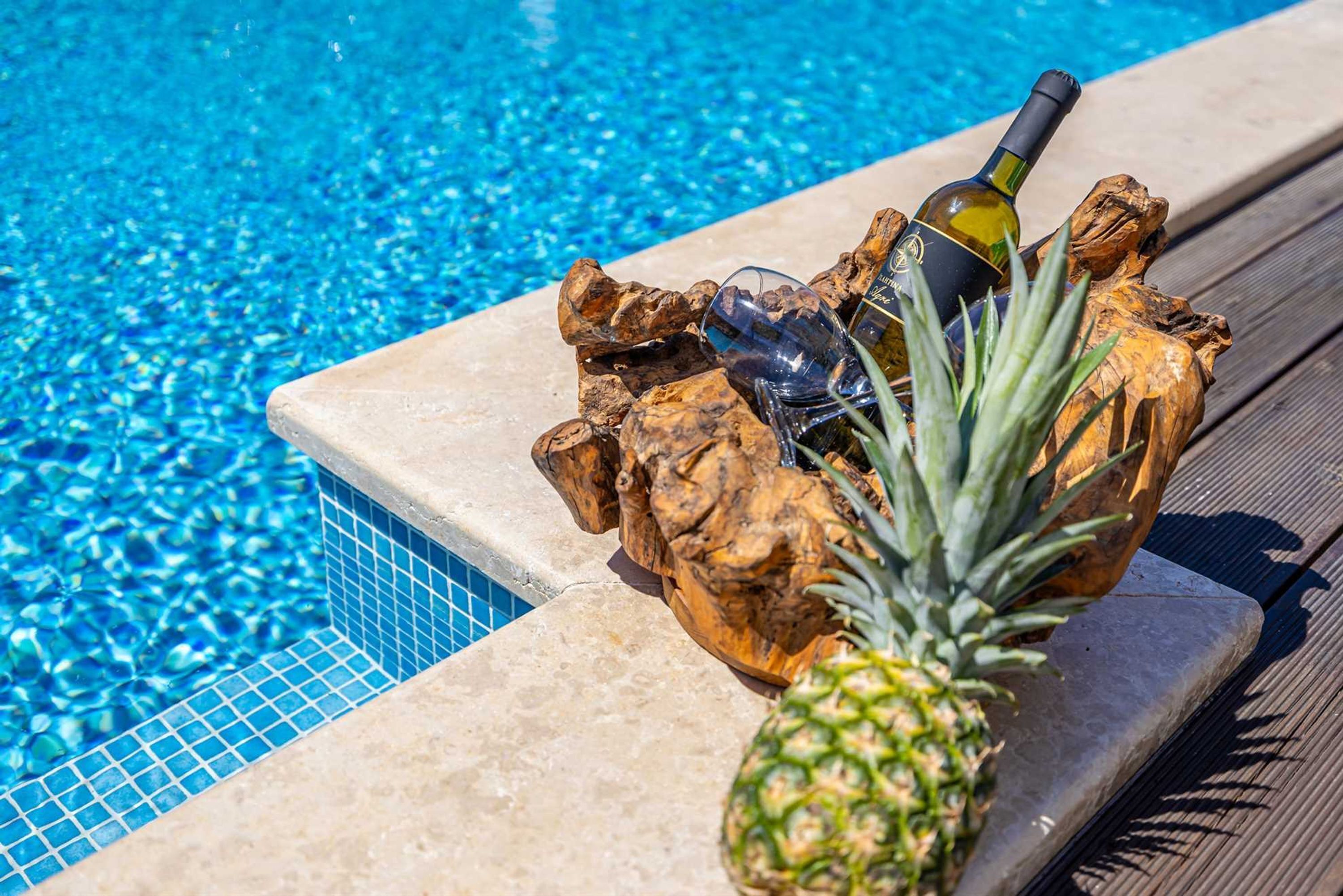 A poolside setting with a decorative wooden bowl containing a bottle, glasses, and a pineapple.