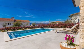 Una piscina con lettini, circondata da un giardino fiorito con fiori rossi e rosa, e case sullo sfondo con tetti in tegole rosse.