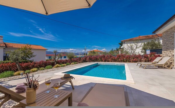 Area piscina con lettini, un tavolo con bevande e vista su case sullo sfondo.