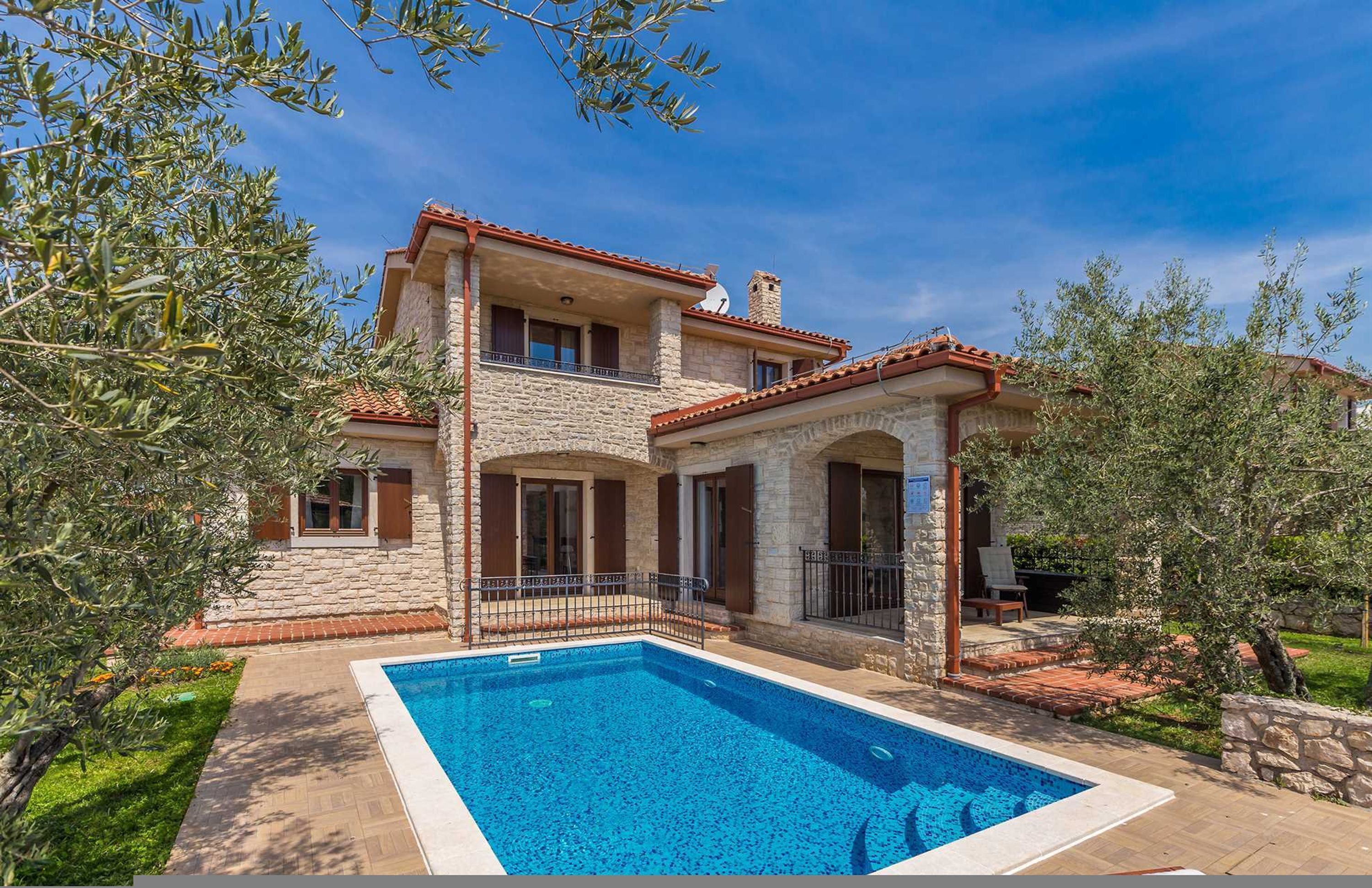 A two-story stone house with a swimming pool, outdoor seating, and olive trees.
