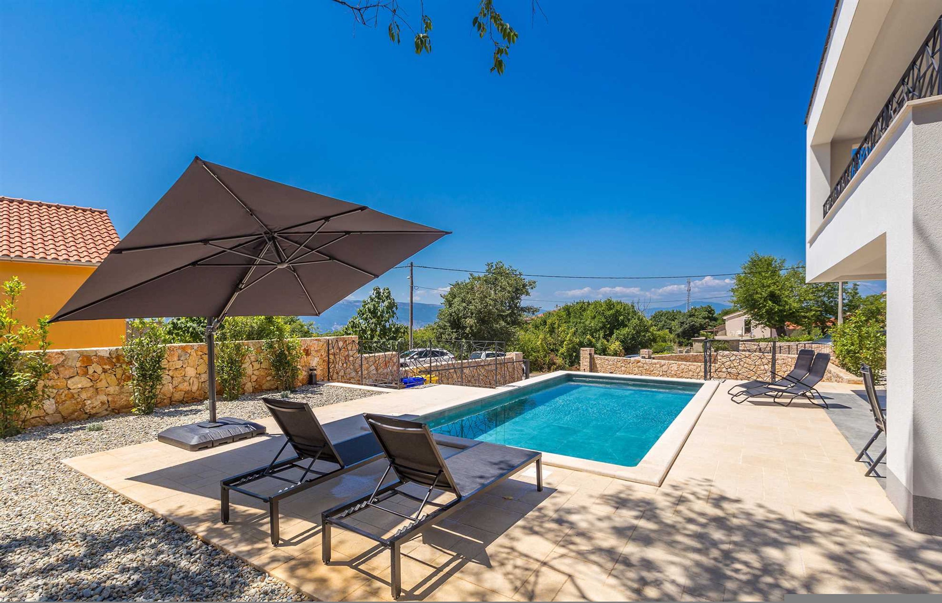 A swimming pool with lounge chairs and an umbrella, surrounded by a stone wall and vegetation.
