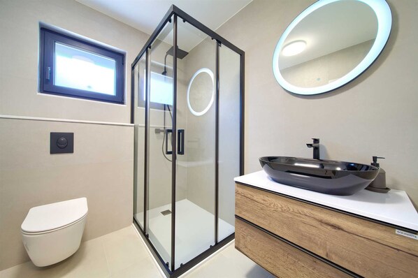 Ein modernes Badezimmer mit einer gläsernen Duschkabine, einer Toilette, einem Waschtisch mit großem Becken und einem Spiegel mit runder Beleuchtung.