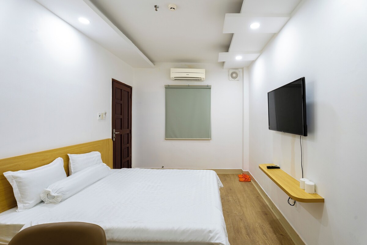 A hotel room with a bed, a TV mounted on the wall, an air conditioning unit, and a wooden shelf with a remote control.