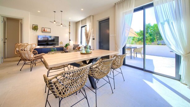 A modern dining area with a wooden table and wicker chairs, a television, and a view of the outdoors through large windows. - Villa Quarnaro With Heated Pool (Omisalj)