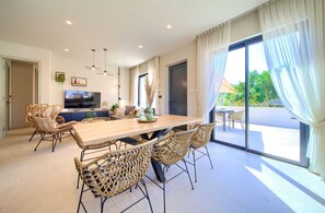 Ein moderner Essbereich mit Holztisch und Korbstühlen, einem Fernseher und Blick ins Freie durch große Fenster.