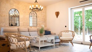 A cozy living room with a stone wall, a chandelier, a sofa, two wicker chairs, and a sliding glass door leading to a garden. - Villa Gita in Malinska (Malinska-Dubasnica)