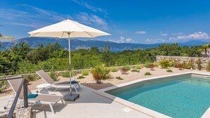 A pool area with lounge chairs and an umbrella, surrounded by a landscaped garden and a view of mountains. - Villa Verena With Heated Pool (Krk)