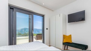 A bedroom with a bed, a bench, a TV, and a sliding glass door leading to a balcony with a view of the sea and mountains. - Villa Verena With Heated Pool (Krk)