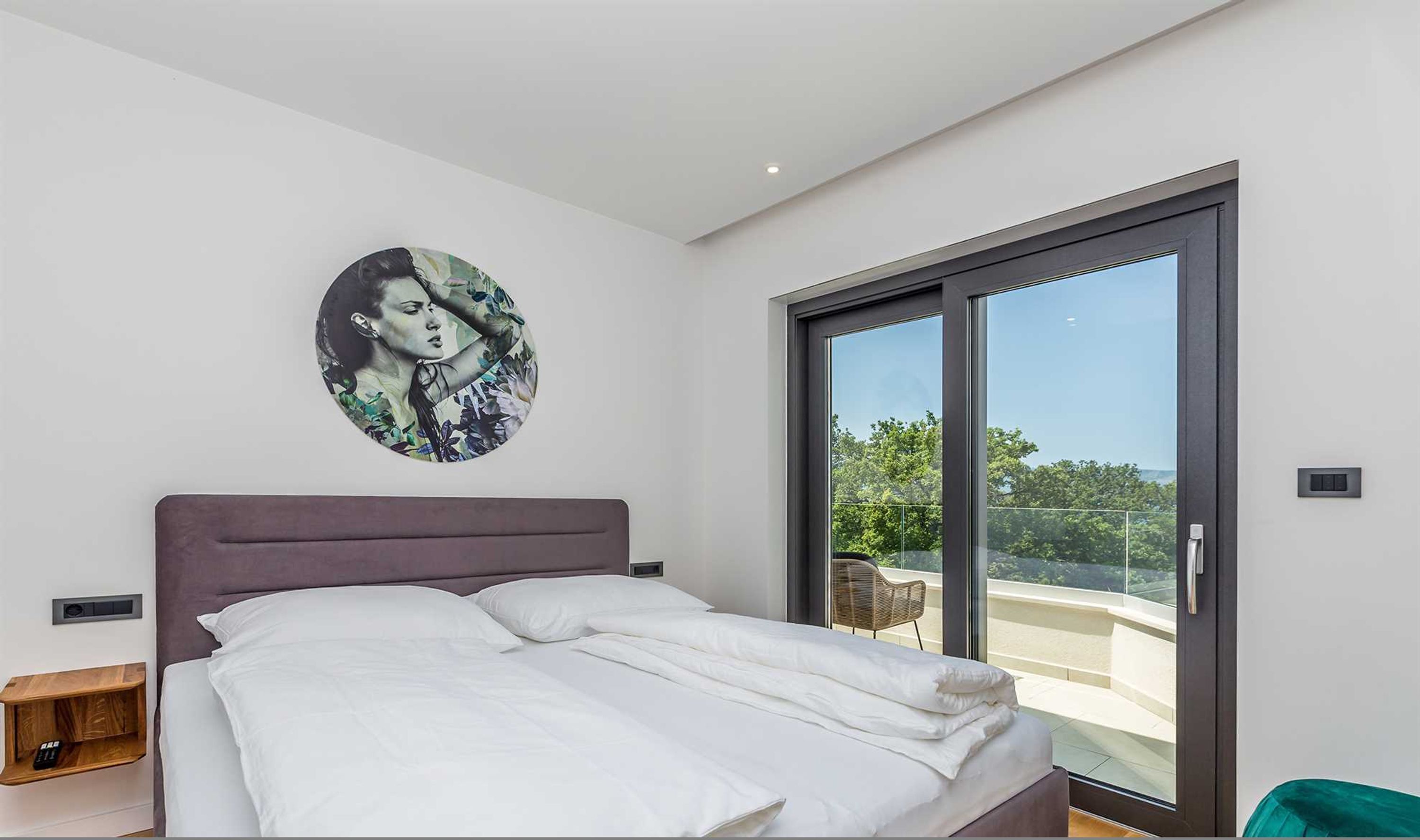 A modern bedroom with a large bed, a circular artwork on the wall, and a sliding glass door leading to a balcony.