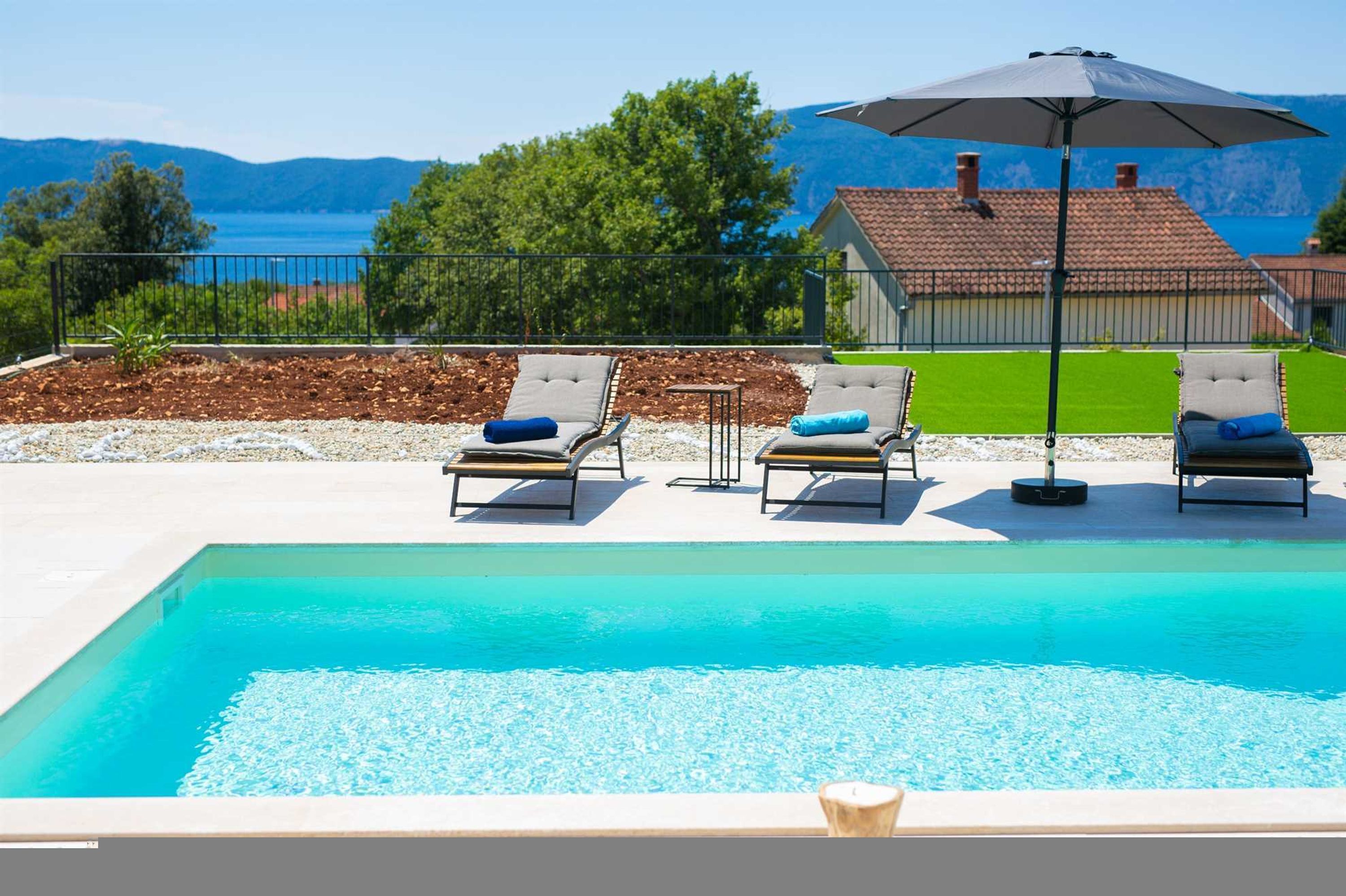 A swimming pool with lounge chairs and an umbrella, surrounded by a landscaped area with a view of a house and mountains.