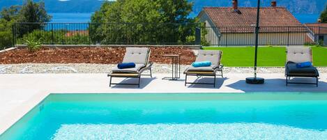 A swimming pool with lounge chairs and an umbrella, surrounded by a landscaped area with a view of a house and mountains.
