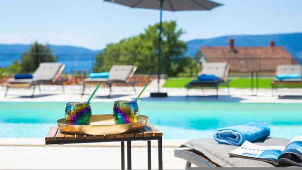 A poolside setting with lounge chairs, a table with two colorful drinks, and a view of a house and mountains in the background. - Villa Magnifica in Pinezici (Krk)