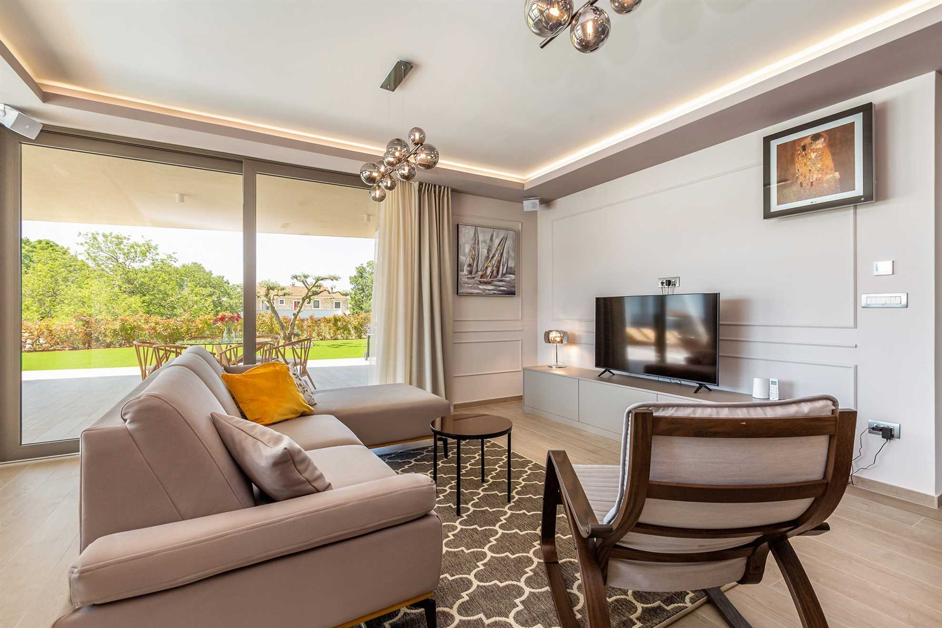 A modern living room with a sofa, a chair, a TV, and a large window offering a view of greenery. - Villa Vista With Private Pool and Sauna (Krk)