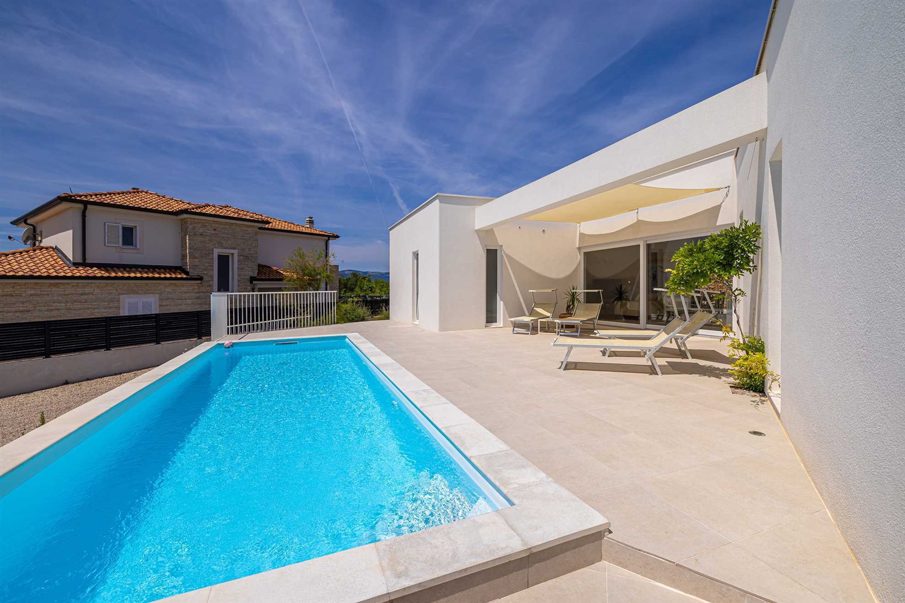 A modern house with a rectangular swimming pool, a covered patio with lounge chairs, and a white exterior wall.