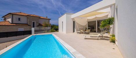 A modern house with a rectangular swimming pool, a covered patio with lounge chairs, and a white exterior wall.