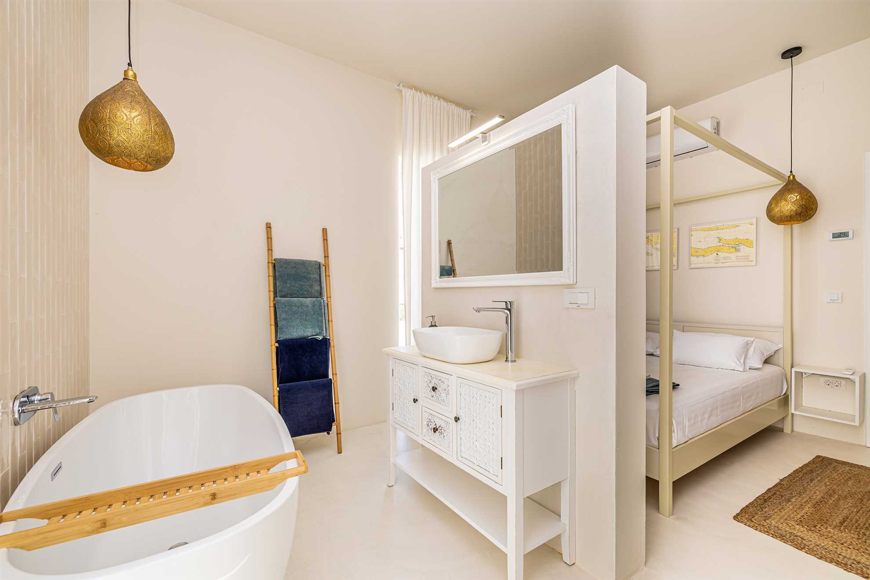 A modern bathroom with a freestanding bathtub, a white vanity with a large mirror, and a bed with a wooden headboard.