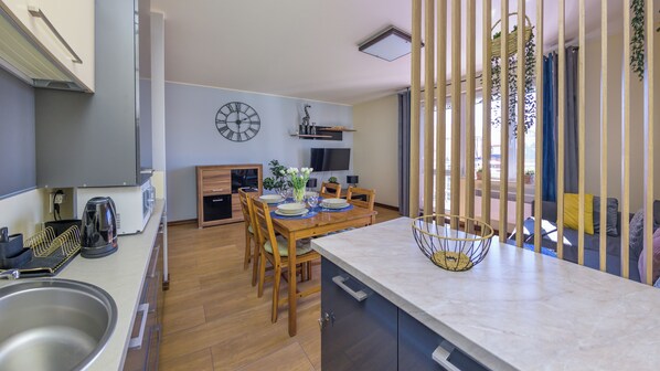 Une cuisine moderne avec des meubles en bois, un coin repas avec une table et des chaises en bois, un téléviseur encastré et une grande fenêtre équipée de stores verticaux.