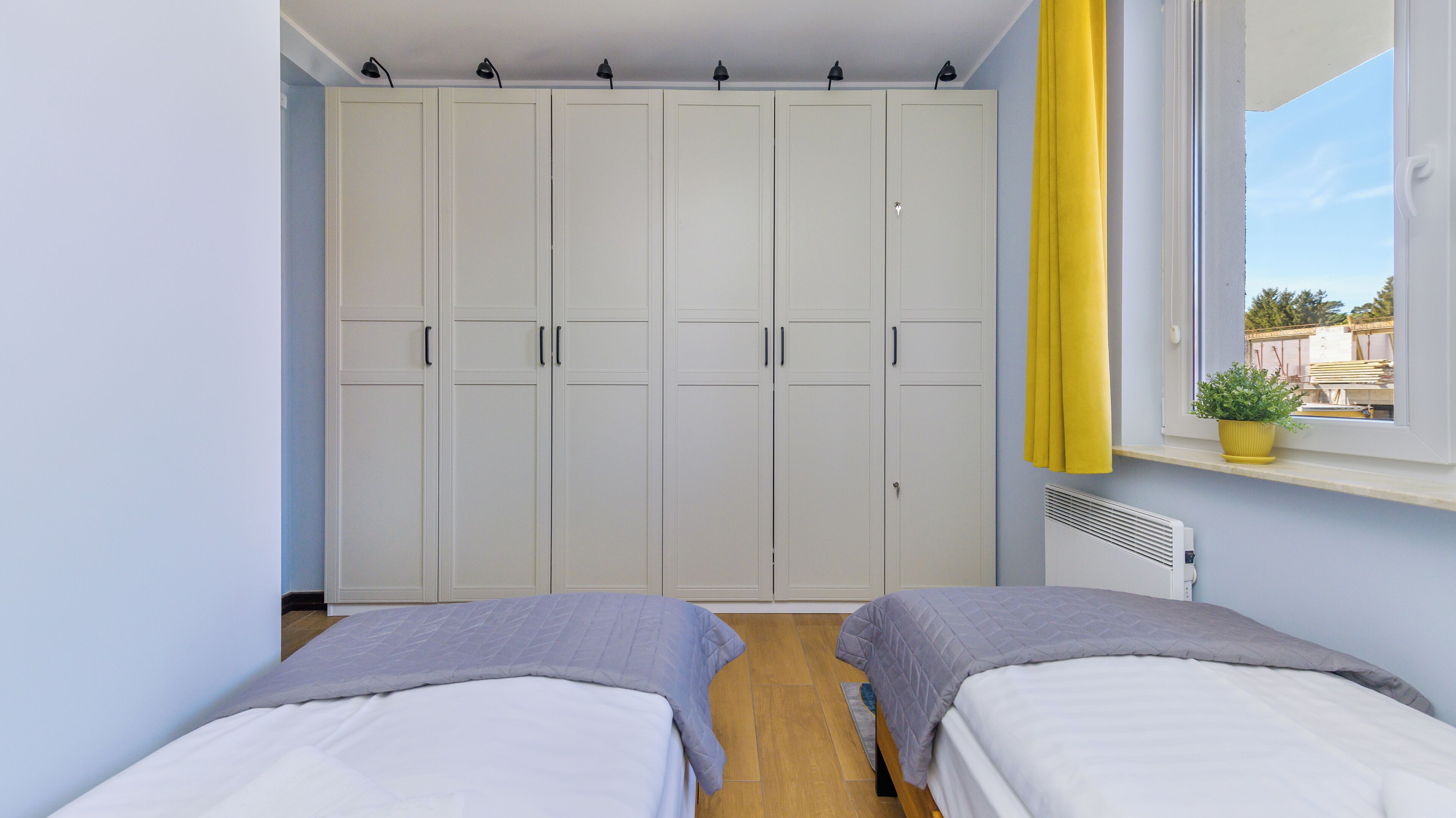 A bedroom with two beds, a wardrobe, a window with yellow curtains, and a potted plant on the windowsill.
