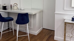 Eine moderne Küche mit weißen Schränken, zwei blauen Barhocker, einer weißen Arbeitsplatte und einem kleinen Tisch mit goldfarbener Rahmen und einem blauen Behälter.