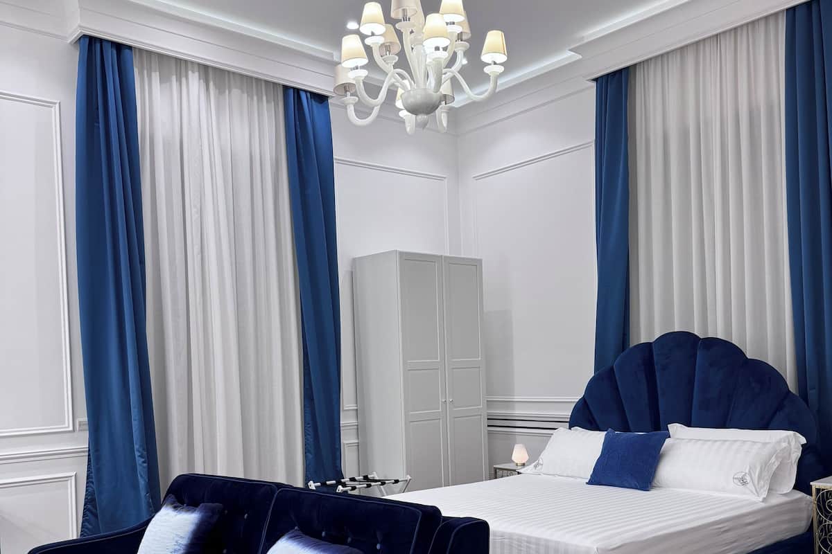 A hotel room with a dark blue sofa, a bed with matching blue bedding, a white wardrobe, and a chandelier.