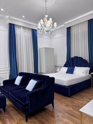 A hotel room with a dark blue sofa, a bed with matching blue bedding, a white wardrobe, and a chandelier.