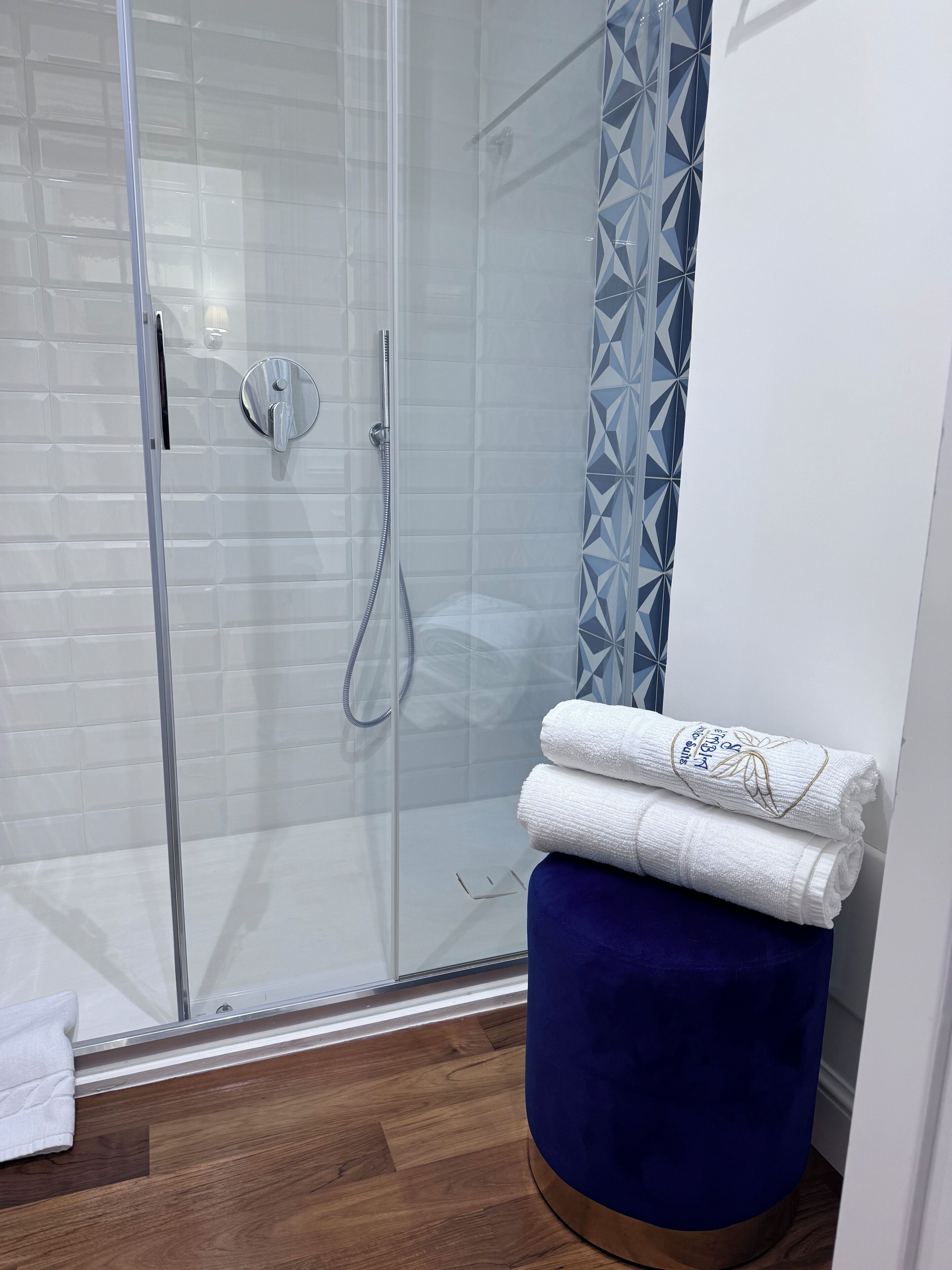 Un baño moderno con cabina de ducha de vidrio, paredes con azulejos blancos y una cortina azul estampada. Hay un taburete azul con una toalla blanca encima.
