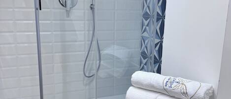Un baño moderno con cabina de ducha de vidrio, paredes con azulejos blancos y una cortina azul estampada. Hay un taburete azul con una toalla blanca encima.