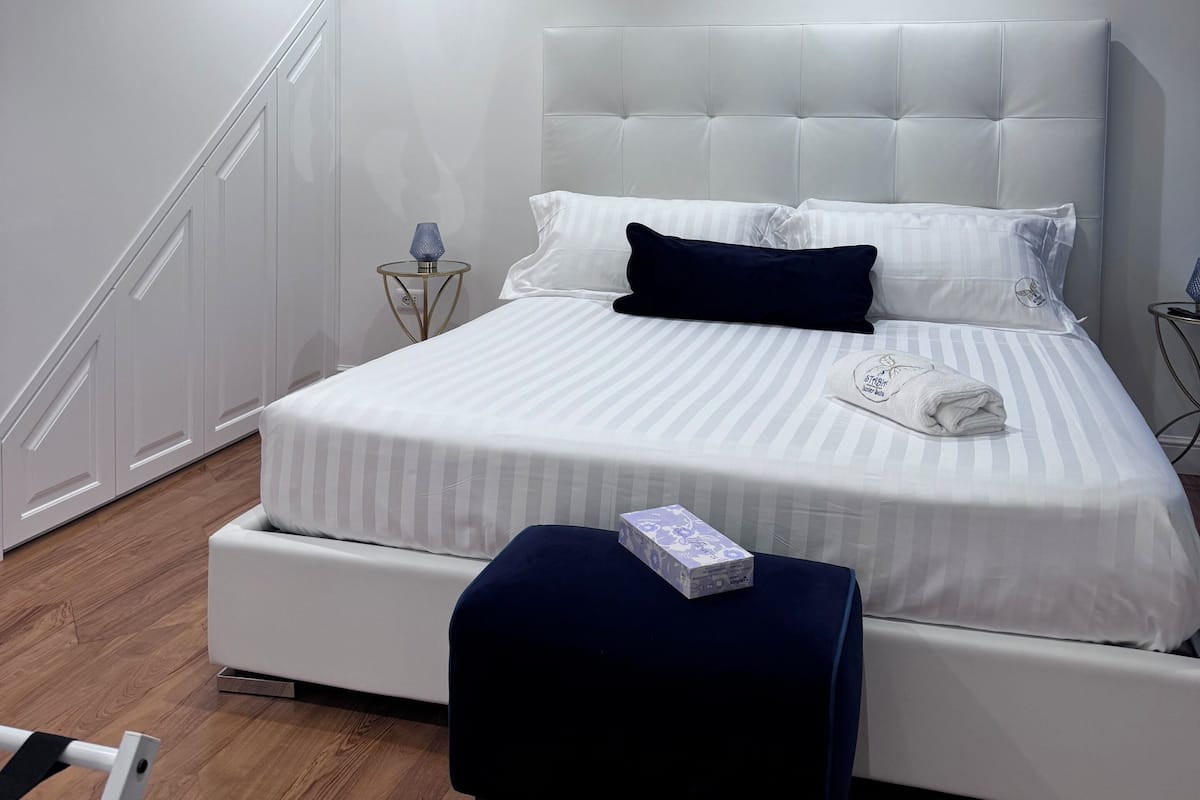 A modern bedroom with a large bed, a bedside table with a lamp, a decorative butterfly on the wall, and a staircase to the left.