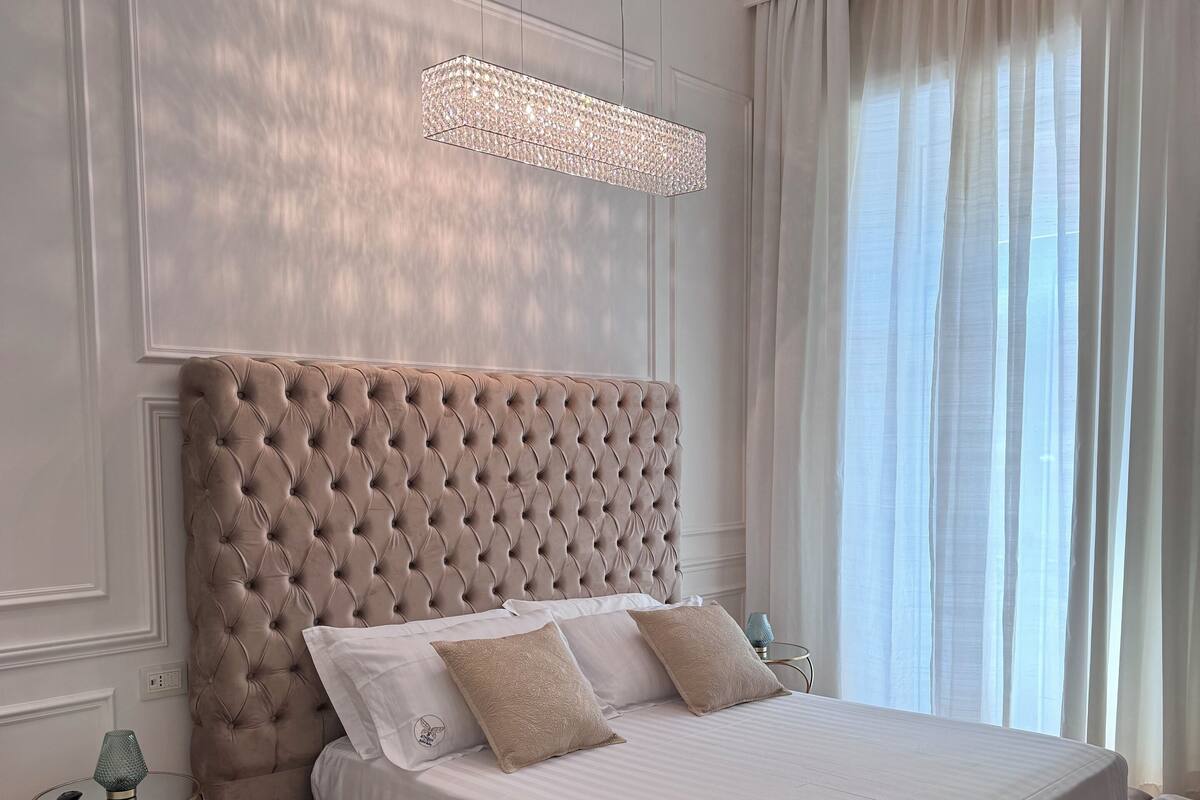 A bedroom with a tufted headboard, a large bed with white linens, and a glass side table with a lamp.