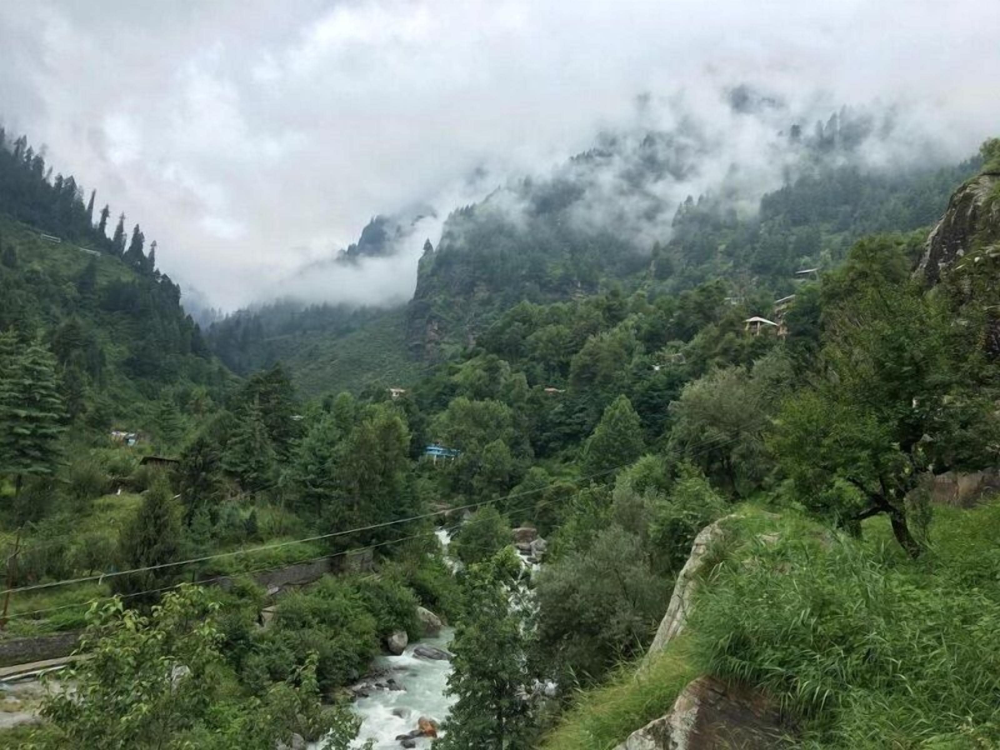 A mountainous landscape with a river, dense greenery, and a few buildings in the distance.