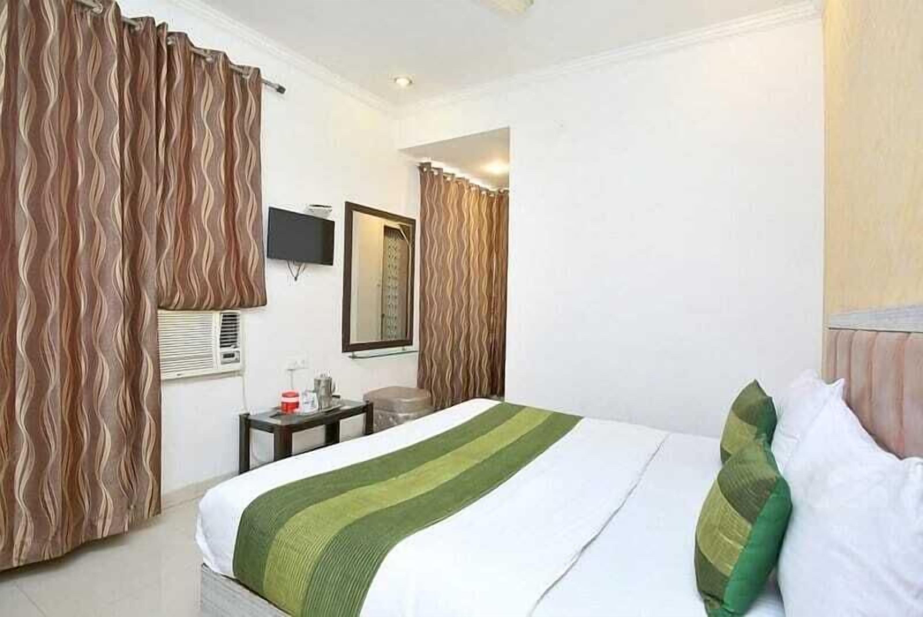 A hotel room with a bed, a television, a mirror, a small table with a red cup, and curtains.