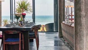 Un área de comedor con vistas al océano, con una mesa dispuesta con platos, vasos y un jarrón de flores.