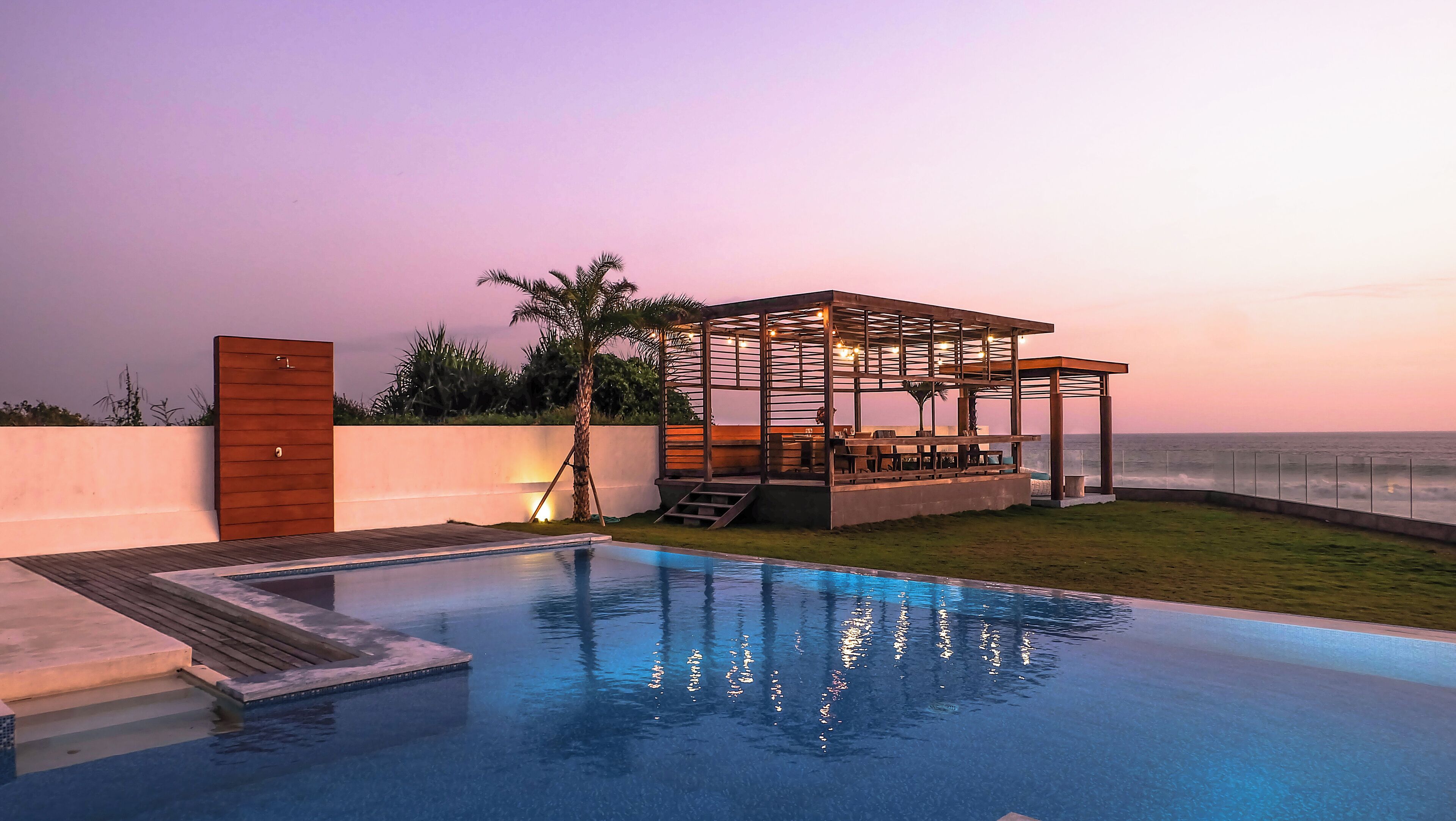 Un espace piscine extérieur moderne, agrémenté d’une pergola en bois et offrant une vue sur l’océan au crépuscule.