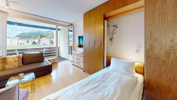 A hotel room with a bed, a sofa, a TV, a wooden wardrobe, and a balcony with a view of buildings and mountains. - Well-kept holiday apartament house in a prime location in St. (St. Moritz)