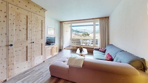 A living room with a sofa, a coffee table, and a TV. There is a large window with a view of buildings and mountains. - Well-kept holiday apartament house in a prime location in St. (St. Moritz)