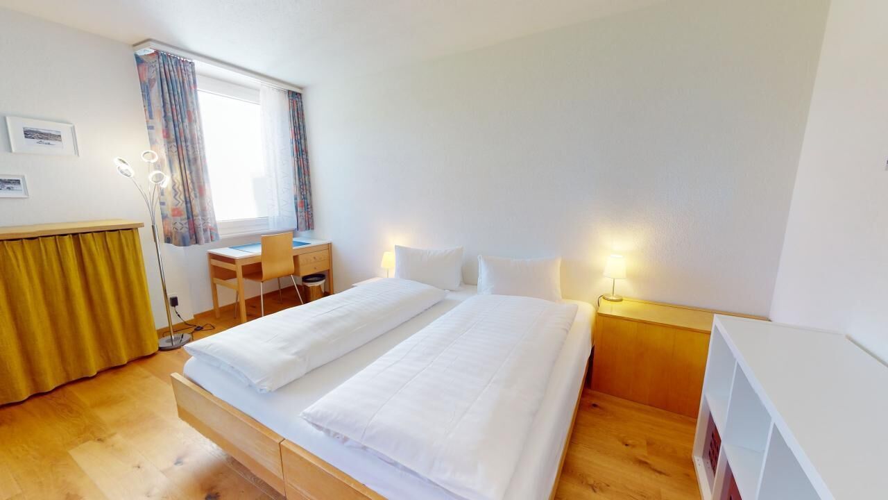 A hotel room with a bed, a desk, a chair, a lamp, and a window with curtains.