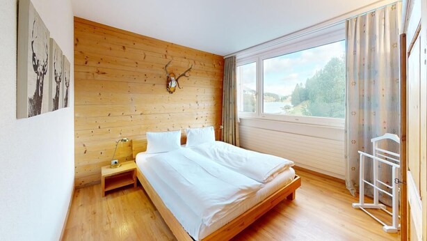 A bedroom with a wooden headboard, a bed with white linens, a nightstand, a window with curtains, and a view of a lake.
