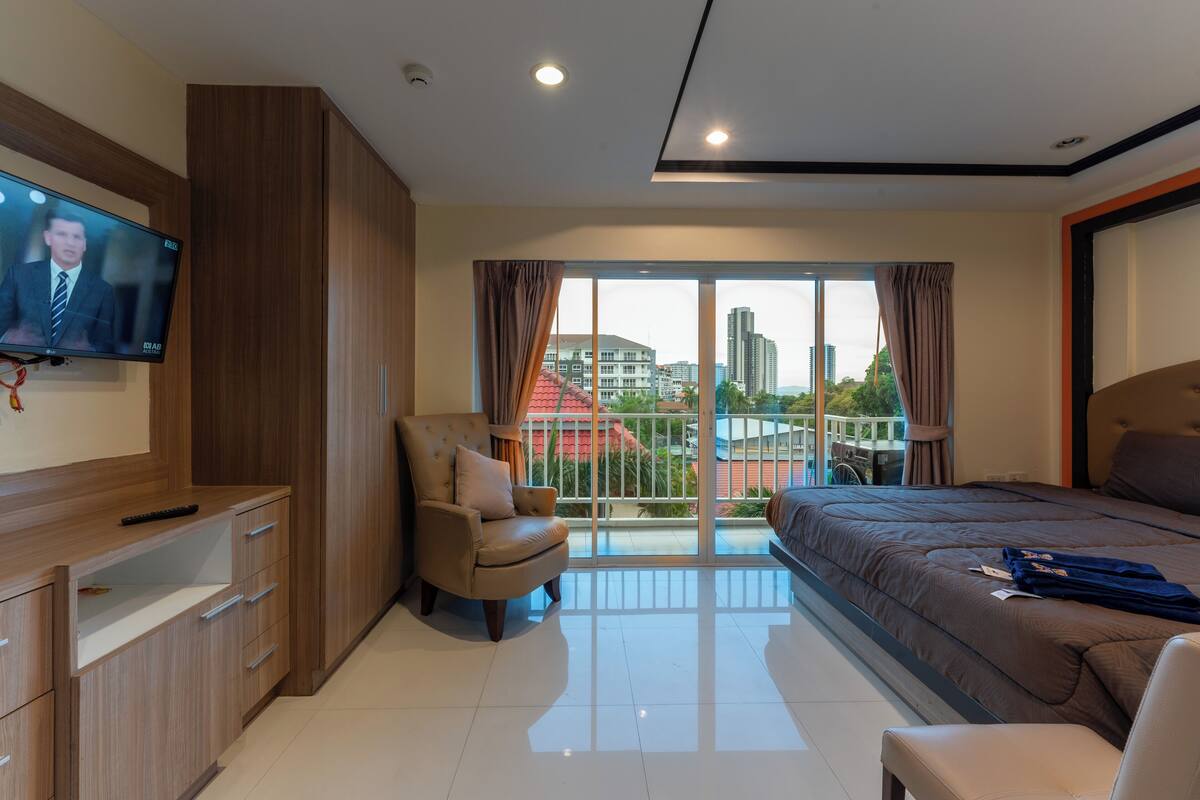 A modern hotel room with a large bed, a TV mounted on the wall, a comfortable armchair, and a balcony with a city view.